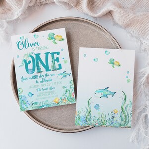 Peut inclure: Une carte d'invitation bleue et verte avec une illustration aquarelle d'une tortue de mer, d'un requin et d'autres cr&eacute;atures marines. Le texte sur la carte indique "Oliver a un an. Rejoignez-nous sous la mer pour c&eacute;l&eacute;brer Samedi 25 avril &agrave; 13h00 Chez les Smith".