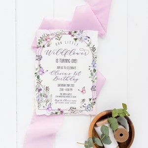 May include: A floral-themed first birthday invitation for Olivia. The invitation features a watercolor illustration of a wreath of flowers and butterflies, with the text "Our Little Wildflower is Turning One!" and details about the party, including the date, time, and location.