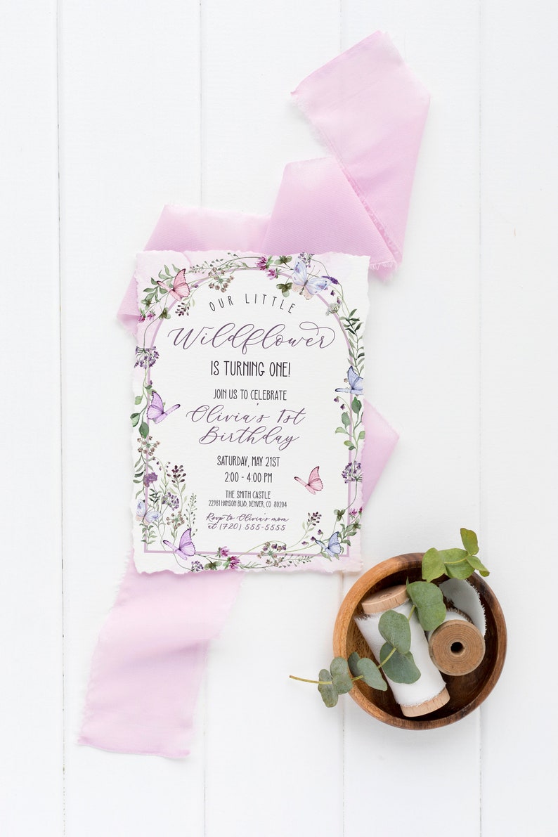 May include: A floral-themed first birthday invitation for Olivia. The invitation features a watercolor illustration of a wreath of flowers and butterflies, with the text "Our Little Wildflower is Turning One!" and details about the party, including the date, time, and location.