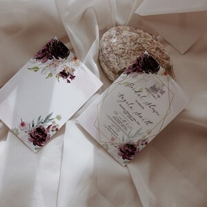 May include: A white card with a floral design in shades of purple and pink. The card is blank and can be used for a variety of purposes. The card is sitting on a white fabric background. The text on the card reads "Bridal Shower for Angela Smith".