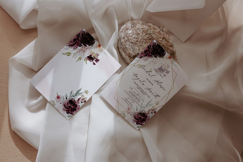 May include: A white card with a floral design in shades of purple and pink. The card is blank and can be used for a variety of purposes. The card is sitting on a white fabric background. The text on the card reads "Bridal Shower for Angela Smith".