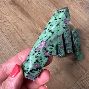 May include: A green, black and pink zoisite crystal point. The crystal is smooth and polished, with a pointed tip. It is a natural stone and has a unique pattern of green, black and pink.