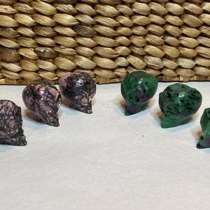 May include: Six small, hand-carved skull-shaped crystal figurines. Three are pink with black veins and three are green with black speckles.