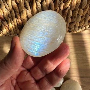 Può includere: Una pietra di luna lucida, di forma ovale, con sfumature iridescenti blu e bianche è tenuta in mano. La superficie della pietra riflette la luce, mostrando un sottile gioco di colori. Ulteriori pietre di luna sono visibili.