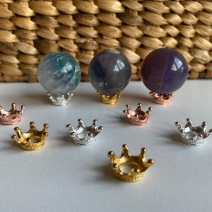 May include: Three small, round, iridescent crystal balls on gold crown-shaped stands. The stands are on a woven wicker surface. There are six additional gold crowns on the surface.