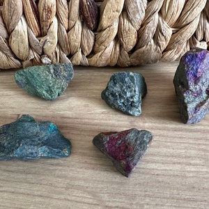 May include: Five rough, irregularly shaped stones with various shades of green, blue, and purple. The stones are arranged on a wooden surface.