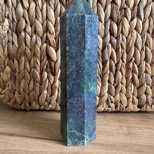 May include: A dark green and black crystal point with a faceted shape. The crystal is sitting on a woven surface.