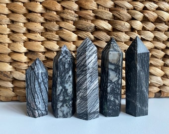 May include: Five black and white zebra jasper crystal points of varying sizes. The points are arranged in a row on a white surface.