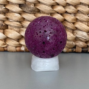 Peut inclure: Une sph&egrave;re de pierre ronde et poreuse de couleur violet fonc&eacute; sur un pi&eacute;destal blanc. La sph&egrave;re a une surface rugueuse et textur&eacute;e.