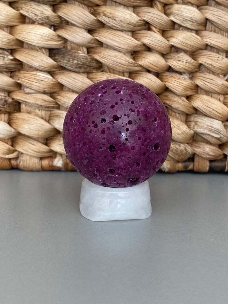 Peut inclure: Une sph&egrave;re de pierre ronde et poreuse de couleur violet fonc&eacute; sur un pi&eacute;destal blanc. La sph&egrave;re a une surface rugueuse et textur&eacute;e.