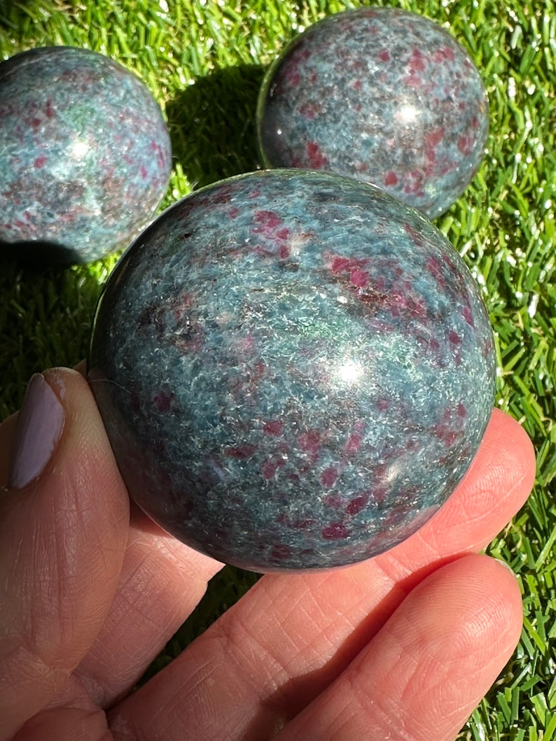 Op de afbeelding: Drie groene en rode edelsteen bollen, &eacute;&eacute;n wordt in een hand gehouden. De bollen zijn gepolijst en hebben een gevlekt uiterlijk.