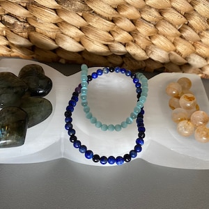 Könnte beinhalten: Eine weiße Selenit-Ladestation mit drei Fächern. Das linke Fach enthält herzförmige Labradorit-Kristalle. Das mittlere Fach enthält zwei Perlenarmbänder, eines blau und eines hellblau. Das rechte Fach enthält kleine, klare Quarzkristalle.