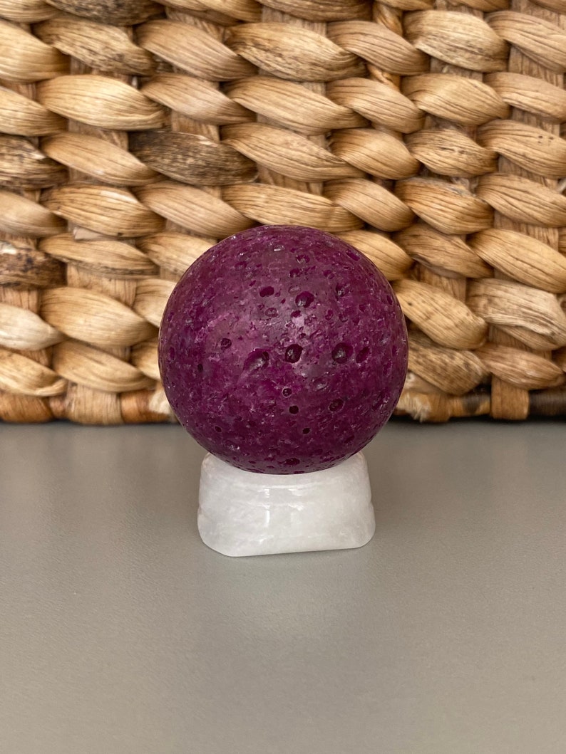Peut inclure: Une sph&egrave;re ronde textur&eacute;e de couleur violet fonc&eacute; sur un pi&eacute;destal blanc. La sph&egrave;re a une surface rugueuse et poreuse.
