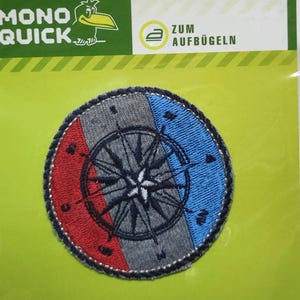 Könnte beinhalten: Ein runder Bügelaufkleber mit einem Kompass-Design. Der Kompass hat einen blau-rot-grauen gestreiften Hintergrund. Der Text "ZUM AUFBÜGELN" ist auf der Verpackung sichtbar.