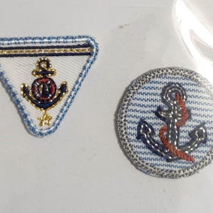 Op de afbeelding: Twee geborduurde strijkbare patches met ankers. Eén patch is een driehoek met een blauw-wit gestreepte rand en een gouden anker met een rode reddingsboei. De andere patch is rond met een blauw-wit gestreepte achtergrond en een blauw anker met een rood touw.