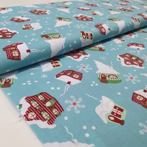 May include: A light blue fabric with a repeating pattern of snow-covered houses in red, green, and brown. White snowflakes and snow drifts are scattered across the fabric. The fabric is folded, showing the texture and pattern.