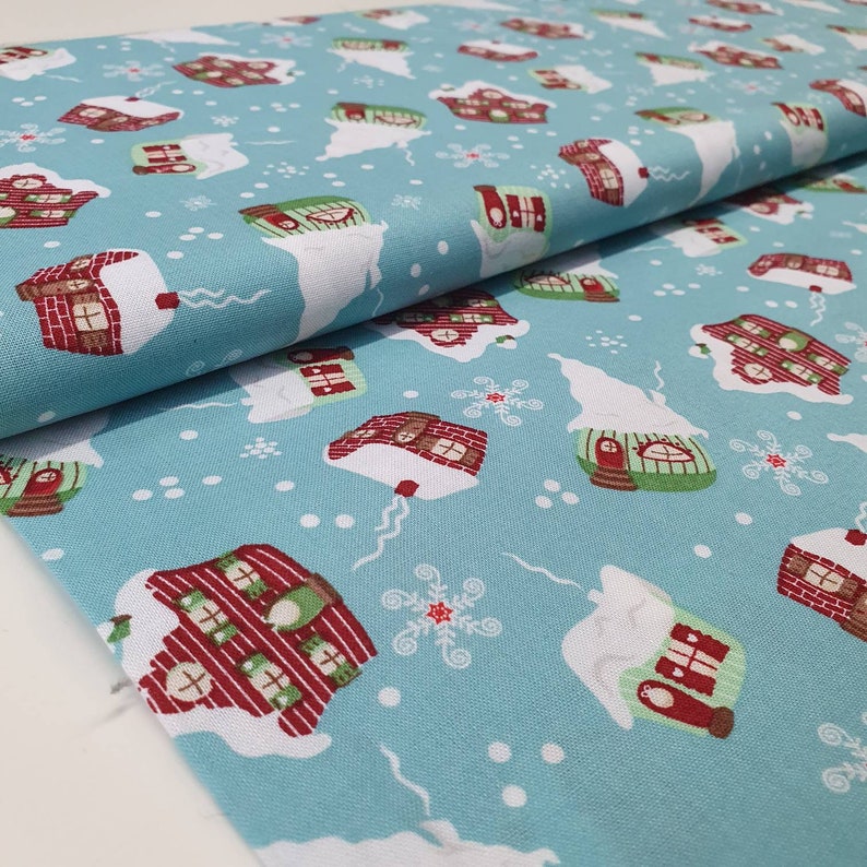 May include: A light blue fabric with a repeating pattern of snow-covered houses in red, green, and brown. White snowflakes and snow drifts are scattered across the fabric. The fabric is folded, showing the texture and pattern.