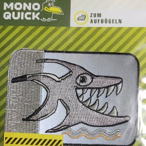 Könnte beinhalten: Ein grauer Bügelaufkleber mit einem Cartoon-Hai mit scharfen Zähnen. Der Aufkleber ist in einer durchsichtigen Plastiktüte mit gelbem und grünem Hintergrund verpackt. Der Text "MONO QUICK" und "REFLEX STICKER SICHERHEIT BEI NACHT!" ist auf der Verpackung sichtbar.