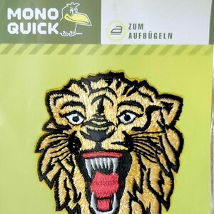 Könnte beinhalten: Bügeleisen-Aufnäher mit einem gelben und schwarzen Tigergesicht mit offenem rotem Maul und weißen Zähnen. Der Aufnäher befindet sich auf einem grünen Hintergrund mit dem Text "MONO QUICK" und "ZUM AUFBÜGELN" in Schwarz.