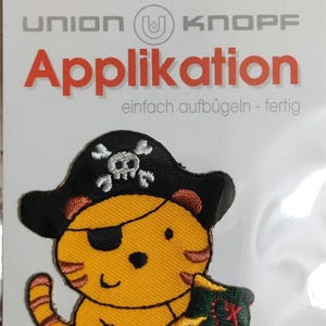 Könnte beinhalten: Bügeleisen-Aufnäher mit einem Cartoon-Tiger, der einen Piratenhut, eine Augenklappe trägt und eine Schatztruhe hält. Der Aufnäher ist gelb, orange, rot, blau, grün und schwarz. Der Text auf der Verpackung lautet "Union Knopf Applikation einfach aufbügeln - fertig".