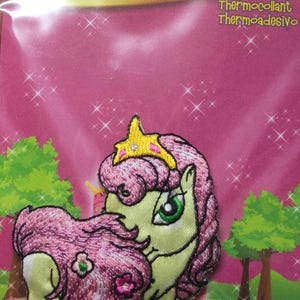Puede incluir: Parche bordado de color rosa y verde para planchar de un pony con una corona y flores en su crin. El texto "Zum Aufbügeln Thermocollant Thermoadesivo" es visible en el embalaje.