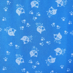 Op de afbeelding: Een blauwe stof met een wit, herhalend patroon van katten met ooglapjes en potafdrukken.