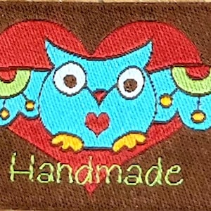 Pode incluir: Uma coruja azul com pés amarelos e um coração vermelho no peito é bordada em um fundo de tecido marrom. A coruja está dentro de um coração vermelho com detalhes verdes e amarelos. O texto "Handmade" é bordado abaixo da coruja.