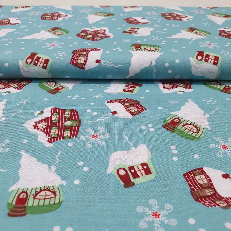 May include: Light blue fabric with a repeating pattern of snow-covered houses in red, green, and white. The houses have chimneys and are surrounded by snowflakes and white snow drifts. A festive, winter-themed textile.
