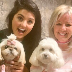 May include: Two women are smiling and holding small white dogs. The dog on the left is wearing a pink sweater and a pink bow. The dog on the right is wearing a pink collar with the word "MUMMY" in white letters.