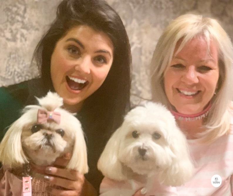 May include: Two women are smiling and holding small white dogs. The dog on the left is wearing a pink sweater and a pink bow. The dog on the right is wearing a pink collar with the word "MUMMY" in white letters.