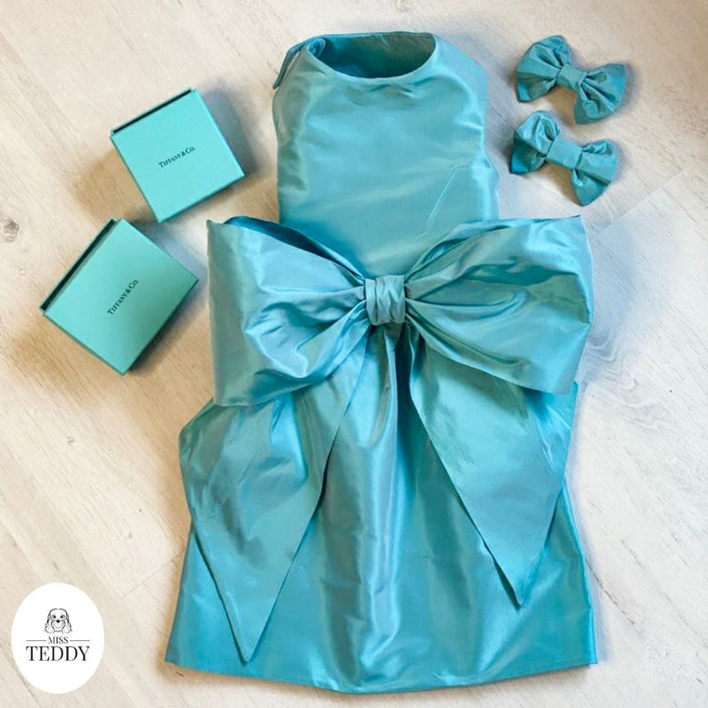 The blue tiffannee dress with swarovski crystals, an oversized bow and two matching hair bows is laid flat on a wooden floor.