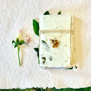 May include: A handmade journal with a bee charm, tied with twine, and surrounded by green leaves on a white background.