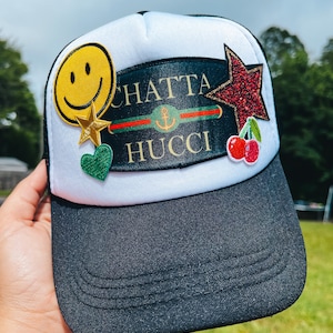 May include: White and black trucker hat with a variety of colorful patches, including a smiley face, a star, a heart, a red star with glitter, a cherry, and a black patch with the text "CHATTA HUCCI" and a green, red, and green stripe.
