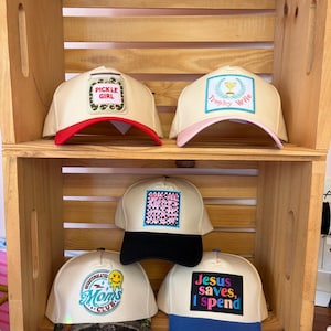 May include: Five baseball caps with cream-colored crowns and varied brim colors. The caps feature embroidered patches with text including Pickle Girl, Trophy Wife, Small Town Nice Show, Overstimulated Moms Club, and Jesus saves, I spend.