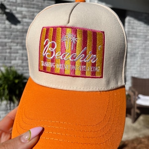 Op de afbeelding: Een beige en oranje baseballpet met het woord "Beachin" geborduurd op de voorkant. De pet heeft een rechthoekige patch met roze en gele strepen en een palmboomontwerp. De tekst "Tanning, Drinking, Relaxing" is ook geborduurd.