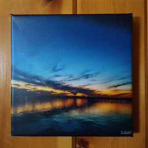 May include: Square canvas art depicting a sunset over water. The sky transitions from deep blue to orange and yellow, reflecting in the water below. A dark silhouette of a bridge or pier is visible. Signed "DANI" in the bottom right corner.