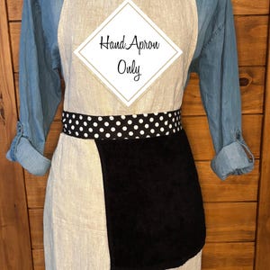 May include: A beige apron with a black towel pocket and a black and white polka dot waistband. The apron has the text "Hand Apron Only" in a white diamond shape. The bottom of the apron has the text "#2-BLACK/POLKADOT".