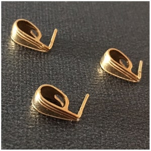 May include: Three gold-colored pendant bails. Each bail has a curved, oval-shaped body with a small loop and a straight, vertical pin extending from the bottom. The bails are on a dark gray surface.