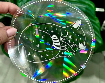 May include: A round, iridescent sticker with a white outline of a sleeping cat with stars and crescent moons. The sticker has a holographic finish.