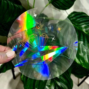 May include: A holographic sticker with a rainbow background and a hand holding three crystals. The crystals are outlined in gold and there are stars and a sun and moon symbol in the background.