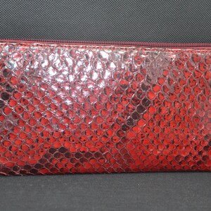 May include: A red, rectangular wallet with a snakeskin pattern. The wallet has a zipper closure and a silver-colored zipper pull. The exterior is a vibrant red with darker red and black accents.