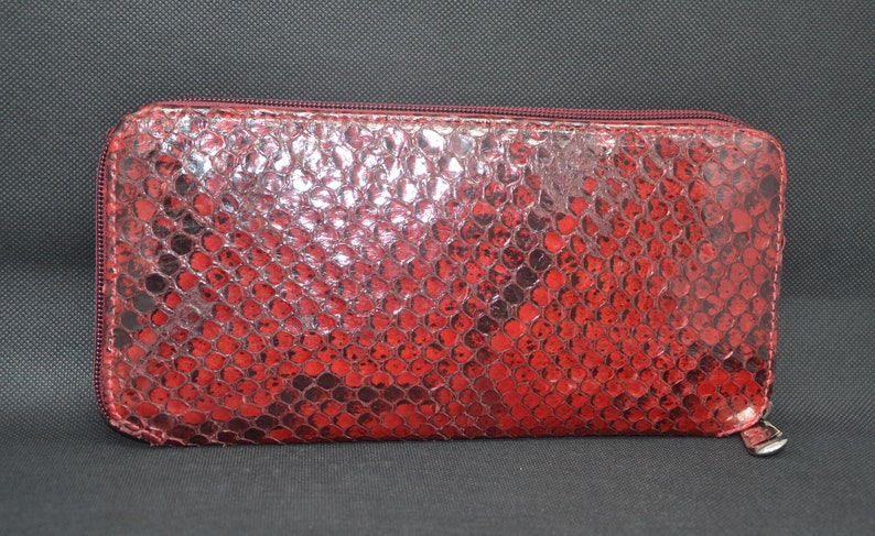 May include: A red, rectangular wallet with a snakeskin pattern. The wallet has a zipper closure and a silver-colored zipper pull. The exterior is a vibrant red with darker red and black accents.