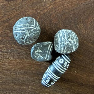 Peut inclure: Quatre perles en pierre gris foncé avec des motifs blancs gravés. Trois perles sont arrondies avec des formes irrégulières et des lignes sculptées, tandis que la quatrième est une perle cylindrique allongée avec un motif de cercles et de bandes.
