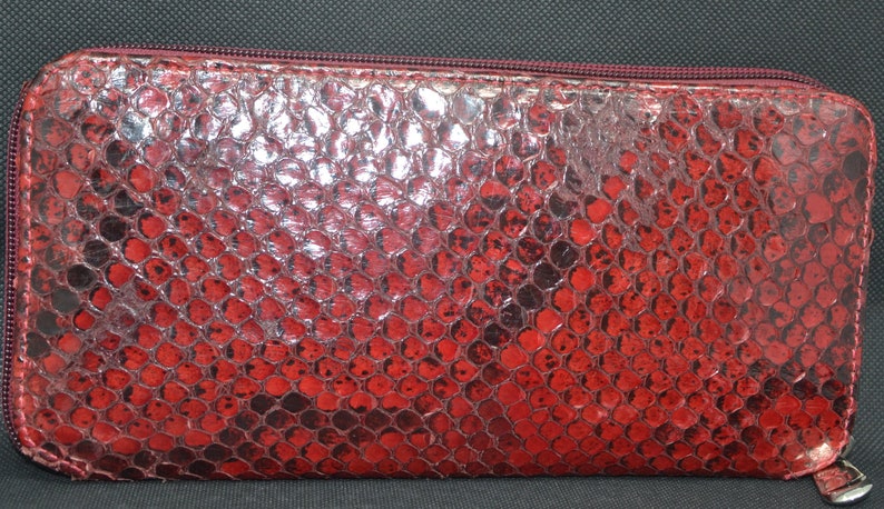 May include: A red, zippered wallet with a snakeskin pattern. The wallet is rectangular and has a burgundy zipper and trim. The snakeskin pattern is a mix of red and black, creating a textured appearance. The wallet is closed.