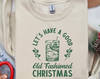 Puede incluir: Una sudadera beige con el texto "Let's have a good Old Fashioned Christmas" en letras verdes. Una ilustración verde de un cóctel old fashioned se encuentra en el centro del texto.