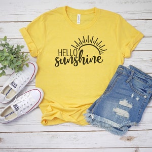 May include: Yellow t-shirt with black text that says "Hello sunshine" and a sun graphic. The shirt is paired with denim shorts and white trainers.
