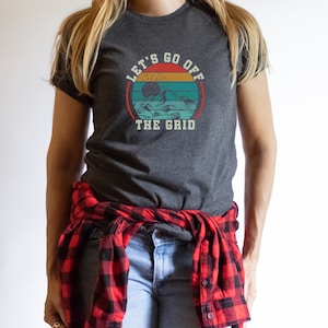 May include: A woman wearing a dark gray t-shirt with a vintage-style graphic that says "Let's Go Off The Grid". The graphic features a sunset with mountains and trees. She is wearing a red and black plaid shirt tied around her waist.
