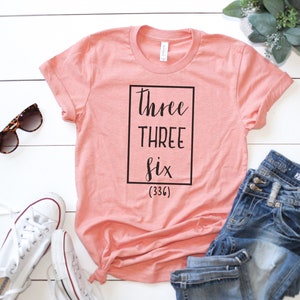 May include: A peach-colored t-shirt with the text "three THREE six (336)" printed in black inside a black square.