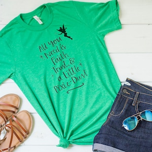 May include: A green t-shirt with the text "All you Need is Faith, Trust, & a Little Pixie-Dust" and a silhouette of a fairy. The shirt is paired with denim shorts, sunglasses, and sandals.