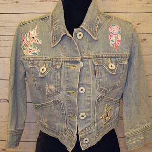 May include: Light blue denim jacket with embroidered unicorn and dreamcatcher patches. Features a cropped fit, button closures, and distressed details. The jacket has two chest pockets and two side pockets. The jacket has a red Levi's tag.